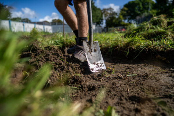 Person digging with a spade with the barbed-wire topped prison wall in the background. (c) Wessex Archaeology