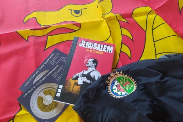 Copy of the play 'Jerusalem' by Jez Butterworth lying on a red flag with a gold wyvern - the flag of Wessex. Also postcards of the tale of the Wiltshire Moonrakers and an item of school uniform with a badge